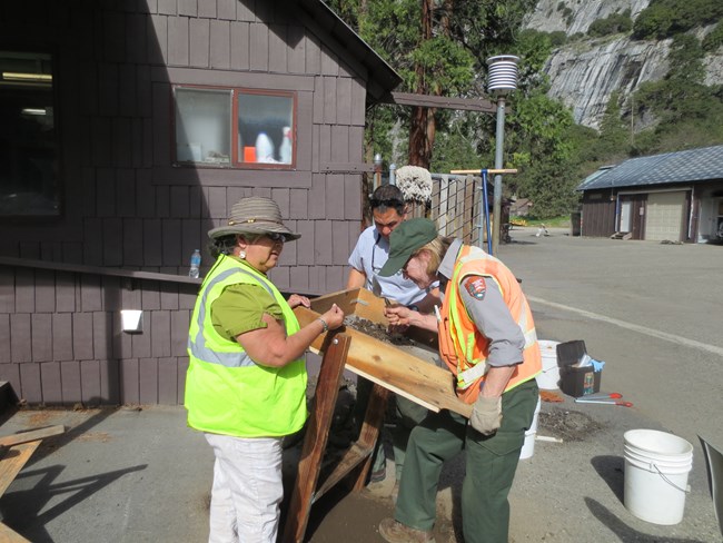 Three people in uniform or hi-vis vests by two rocking archeological sifters. The two people on the right are using the sifters while the person on the left is examining an item.