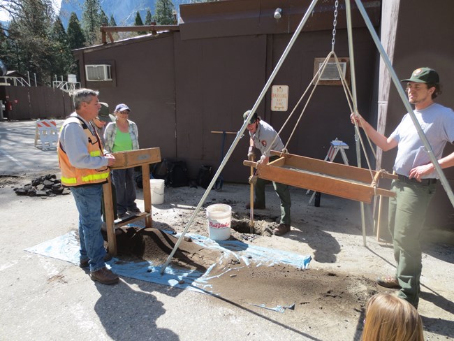 A group of people in NPS uniform or hi-vis vests stand around a hanging archeological sifting frame. Others stand on the side by a rocking sifting screen.