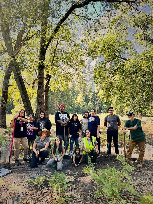 A picture of a group of seventeen Tribal members. They stand with rakes and other tools in front of black oak trees.