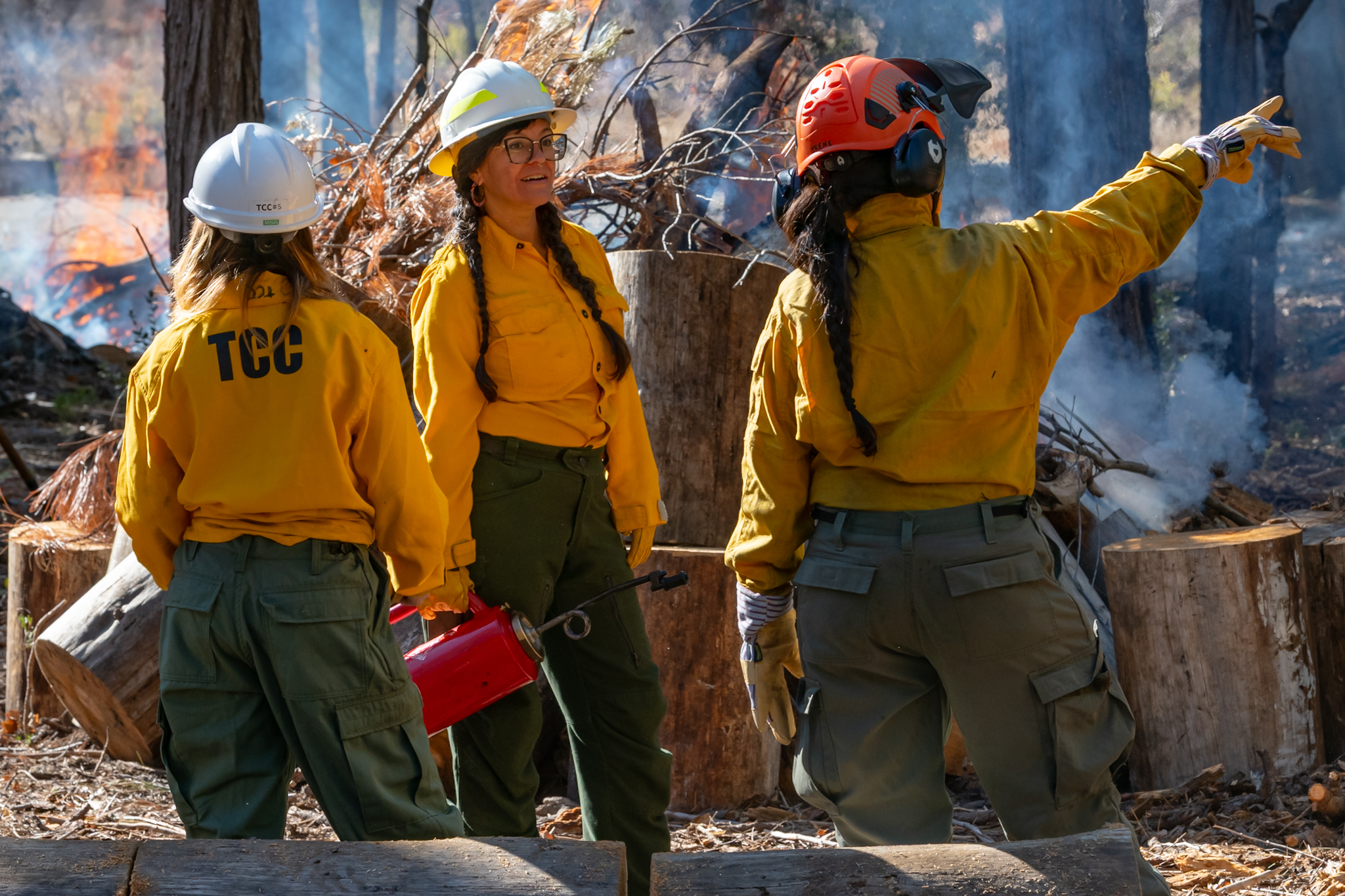 Three women in yellow jackets, cargo pants, and hard hats stand in front of a fire. One holds a fuel torch. They are talking or pointing toward something offscreen.