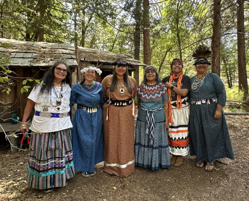 Six women wearing traditional Tribal regalia pose for a picture in front of a cabin and trees.