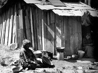 A black and white historical image of two Native women sitting by a cabin in Yosemite Valley. One woman sits in the shade of a doorway, and the other woman sits in front of the cabin with a woven basket and other items.