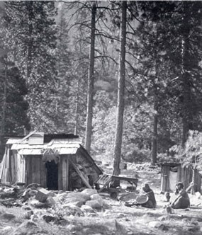 Two American Indians sit on ground by their simple housing