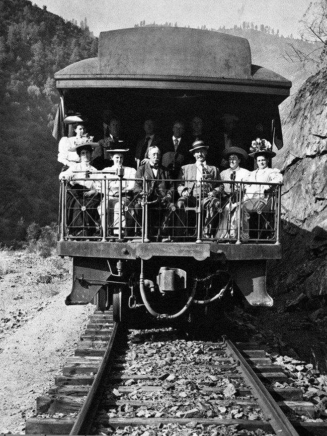 A black and white image of a group of people in early 20th century clothes pose in the back of an open air railcar