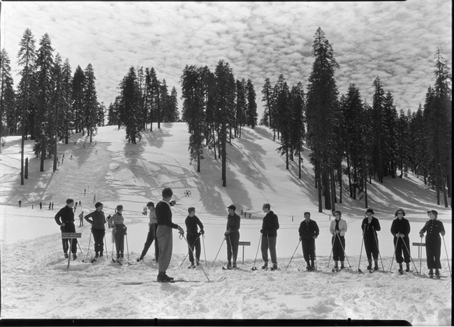 A row of skiiers face forward in front of a snow covered hill with more skiiers. In front of them, a man points to the hill with a ski pole.