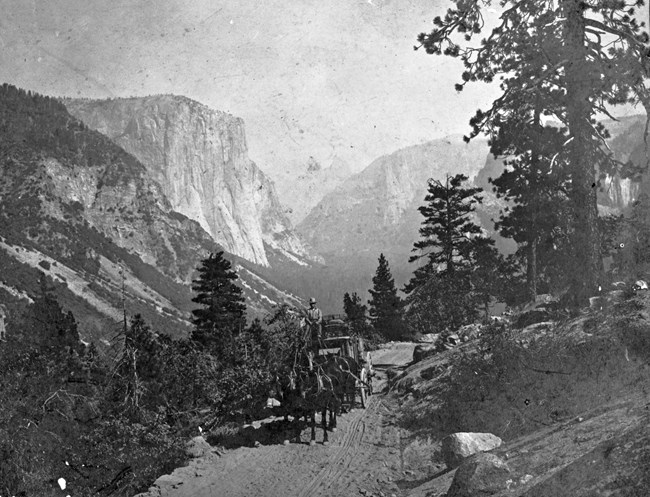 Four horses and a wagon travel along an unpaved road with spectacular granite peaks behind them