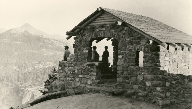 A man in a hut comprised of stone and wooden logs looks out over the distant landscape.