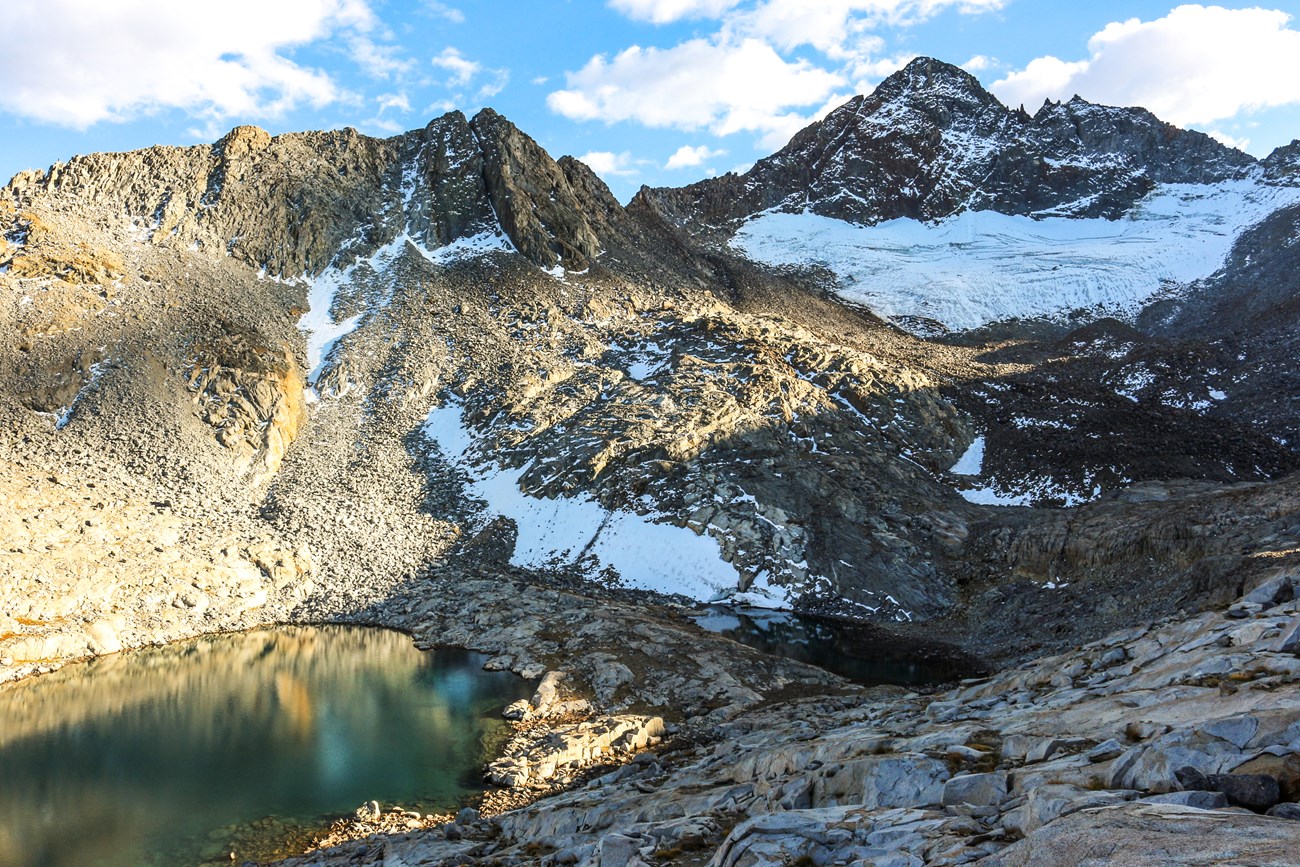 Photo of mountain peaks with a small glacier under one peak and snow scattered elsewhere, and a lake