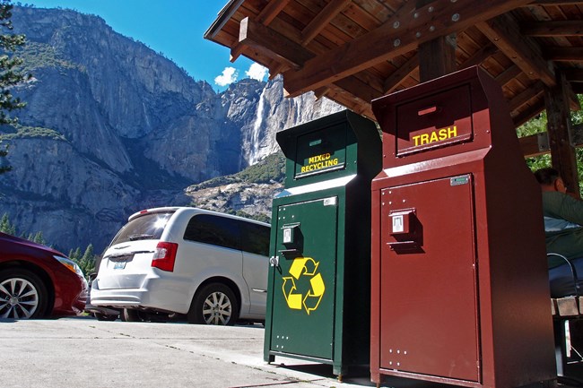 A brown trash can and a green recycling bin stand in the foreground, with large waterfall visible in background.