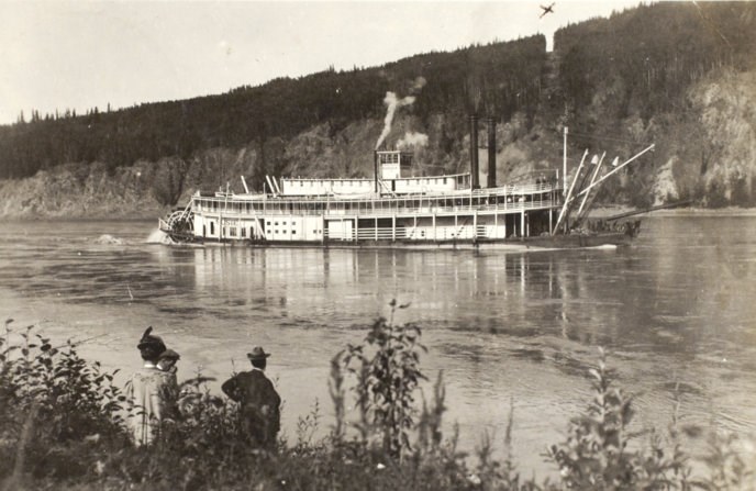 Historic photo of a riverboat on the Yukon River passing in front of the new international boundary which is a bare swath cut through the trees. Historic photo of a riverboat on the Yukon River passing in front of the new international boundary which is a bare swath cut through the trees.
