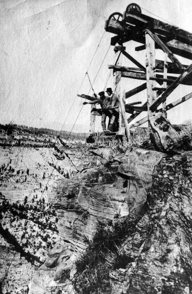 Black and white photo of two men standing on the edge of Zion Canyon with a wooden structure behind them with wires extending down into the canyon.