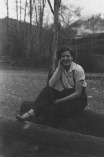 Young woman sitting on logs, wearing a white button down, black pants, and black suspenders