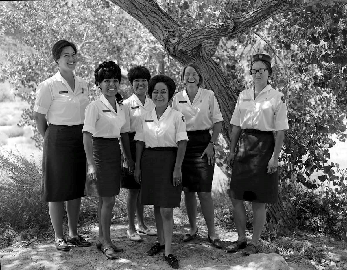 Six uniformed women, in white shirts and black skirts, standing together, smiling at the photographer
