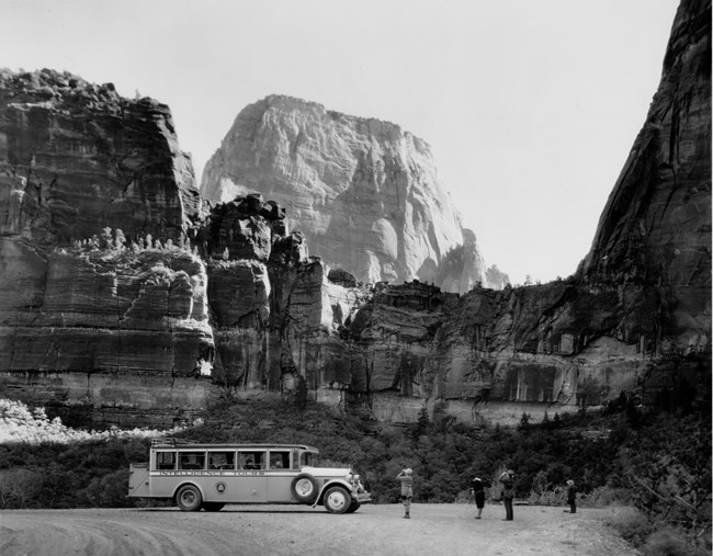 Historic photo of steep rocks above visitors standing near a tour bus in a parking lot.