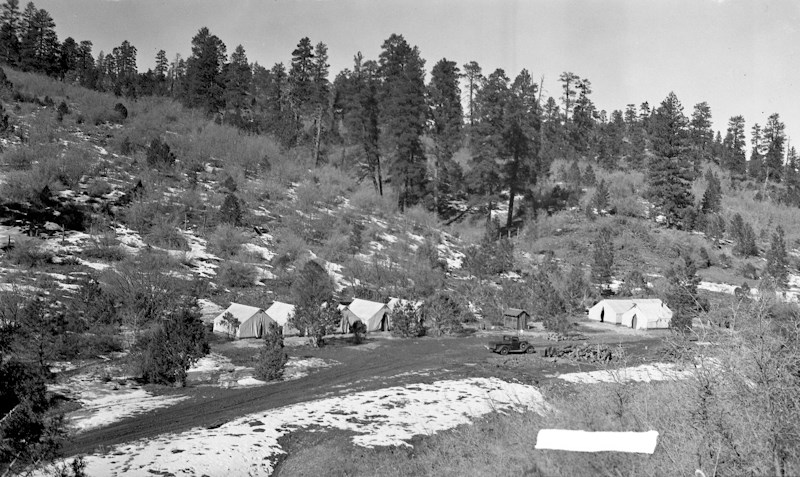 Black and white photo of white tents with trees in the background, patches of snow is on the ground.
