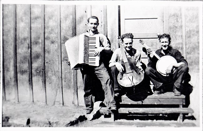 Three men sit on a step outside a building and play an accordion, guitar, and banjo.