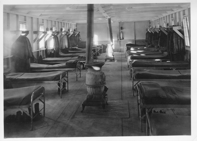 Black and white photo of the interior of a building, a large room with many cots lining the walls, there is a wood stove in the center for heat.
