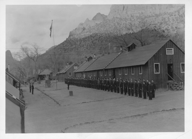 ZION 7097E: After breakfast, the men would stand in attention in front of the colors during the raising of the flag before heading off to work. This photo shows the enrollees standing at attention in front of the flagpole at Bridge Mountain Camp in Zion.