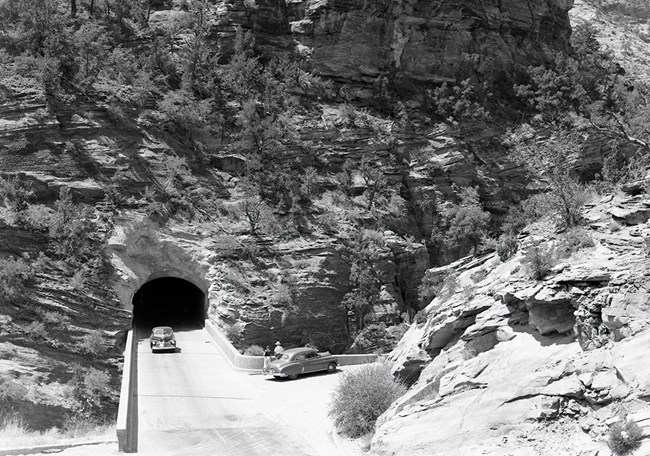 Car drives out of tunnel into an area surrounded by high rocks.