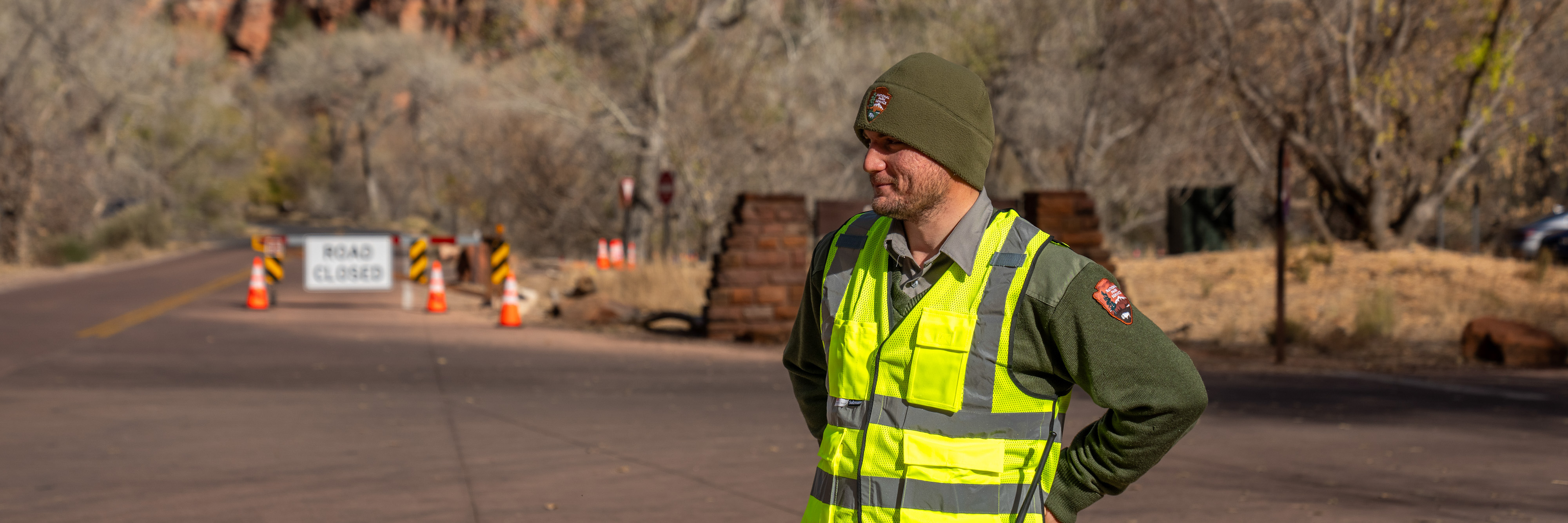 A park ranger in a high-visibility vest stands along a roadway.