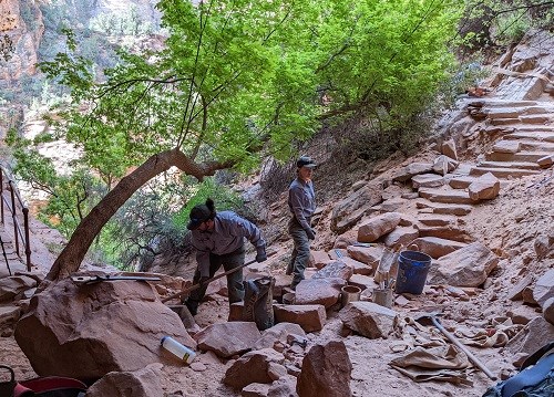 To staff members work on the Canyon Overlook trail surrounded by several rocks being prepared for use in trail repairs.