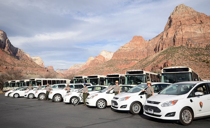 Rangers stand with hybrid and plug-in hybrid vehicles near Zion Canyon Shuttle Line propane powered shuttles with the walls of Zion Canyon behind them.