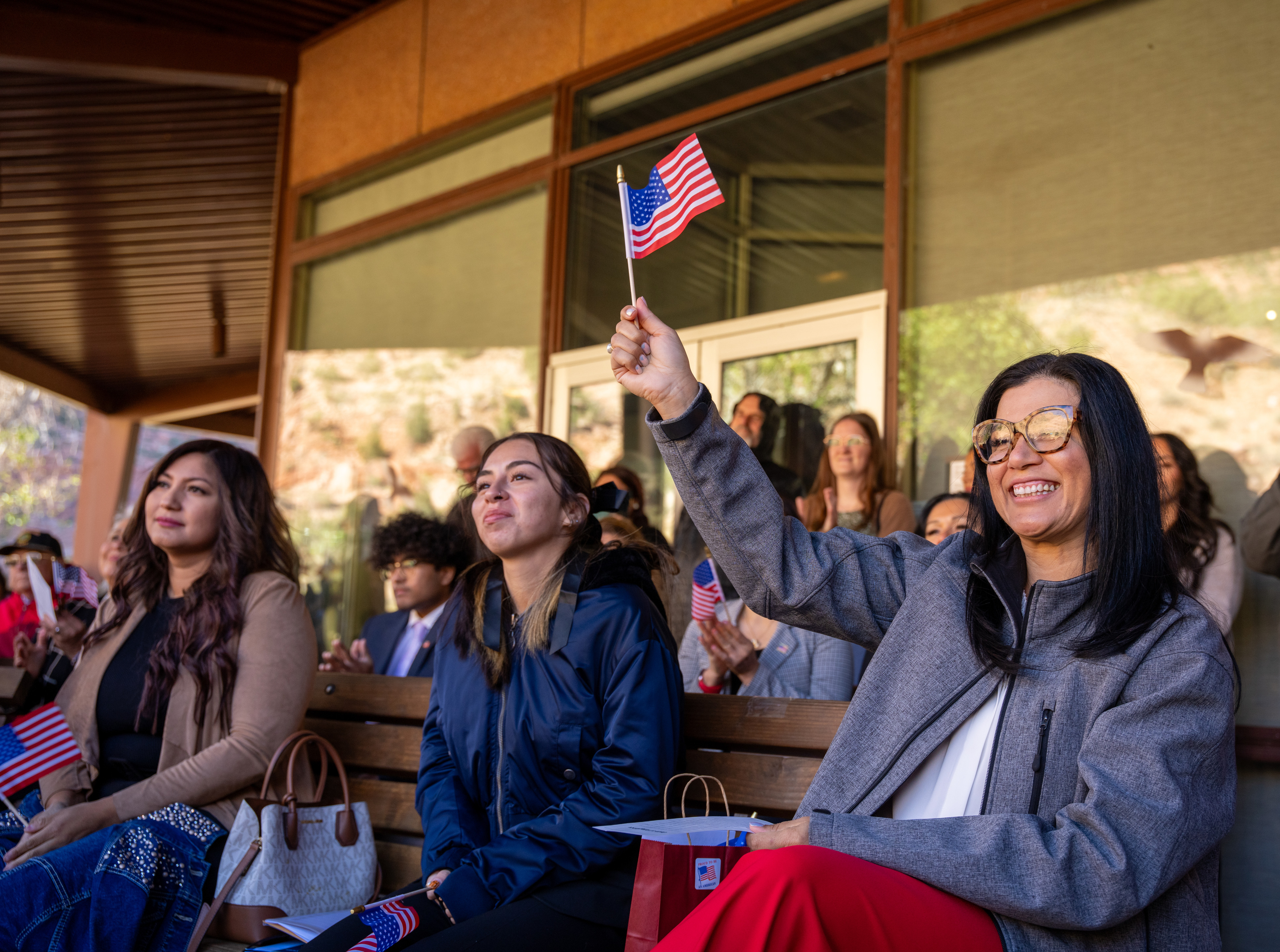 2024 Zion National Park Naturalization ceremony A woman sitting on a bench amongst a crowd smiling and waving an American Flag