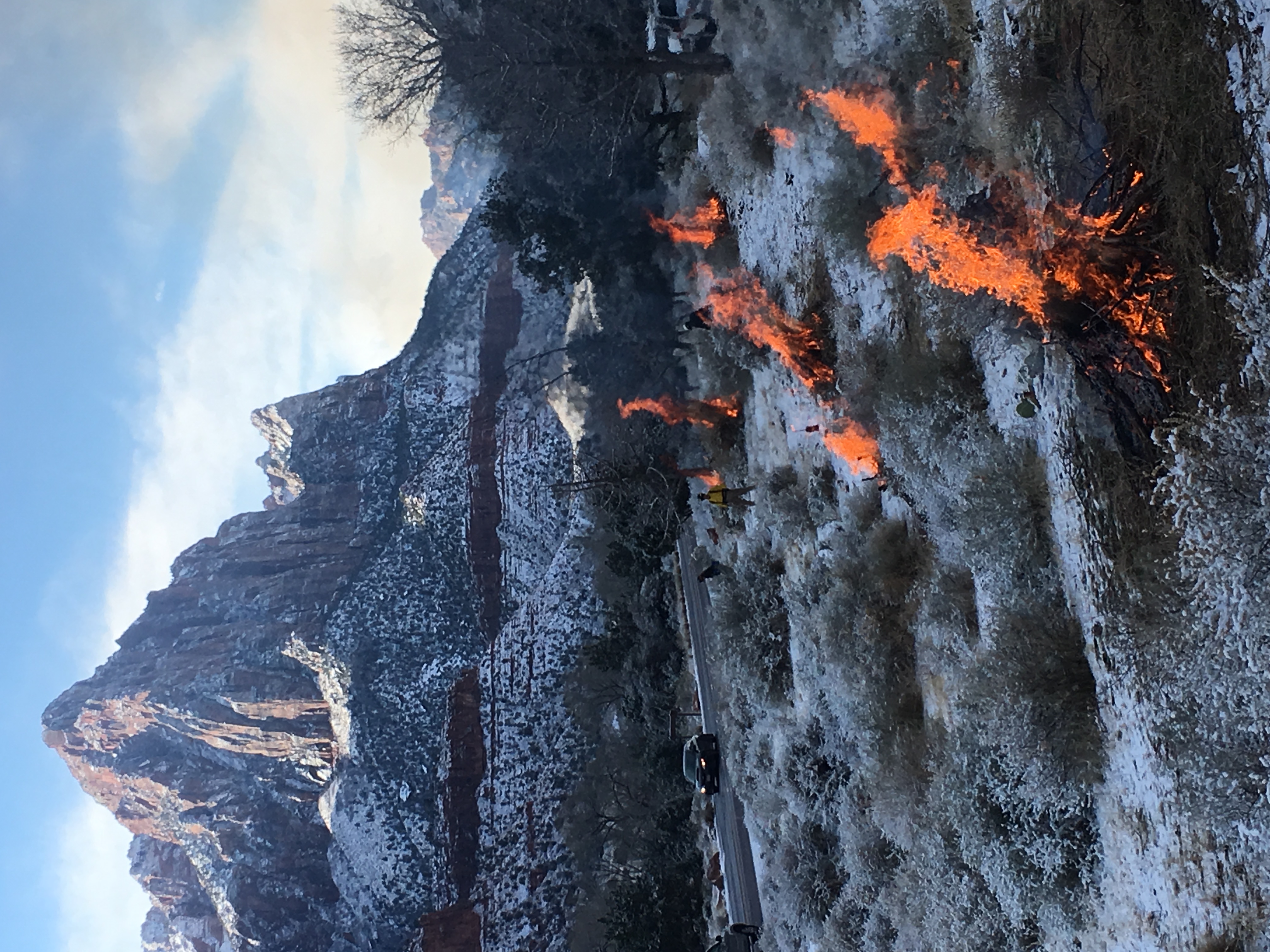 Several piles of debris are on fire in a snowy landscape with Watchman Peak in the background