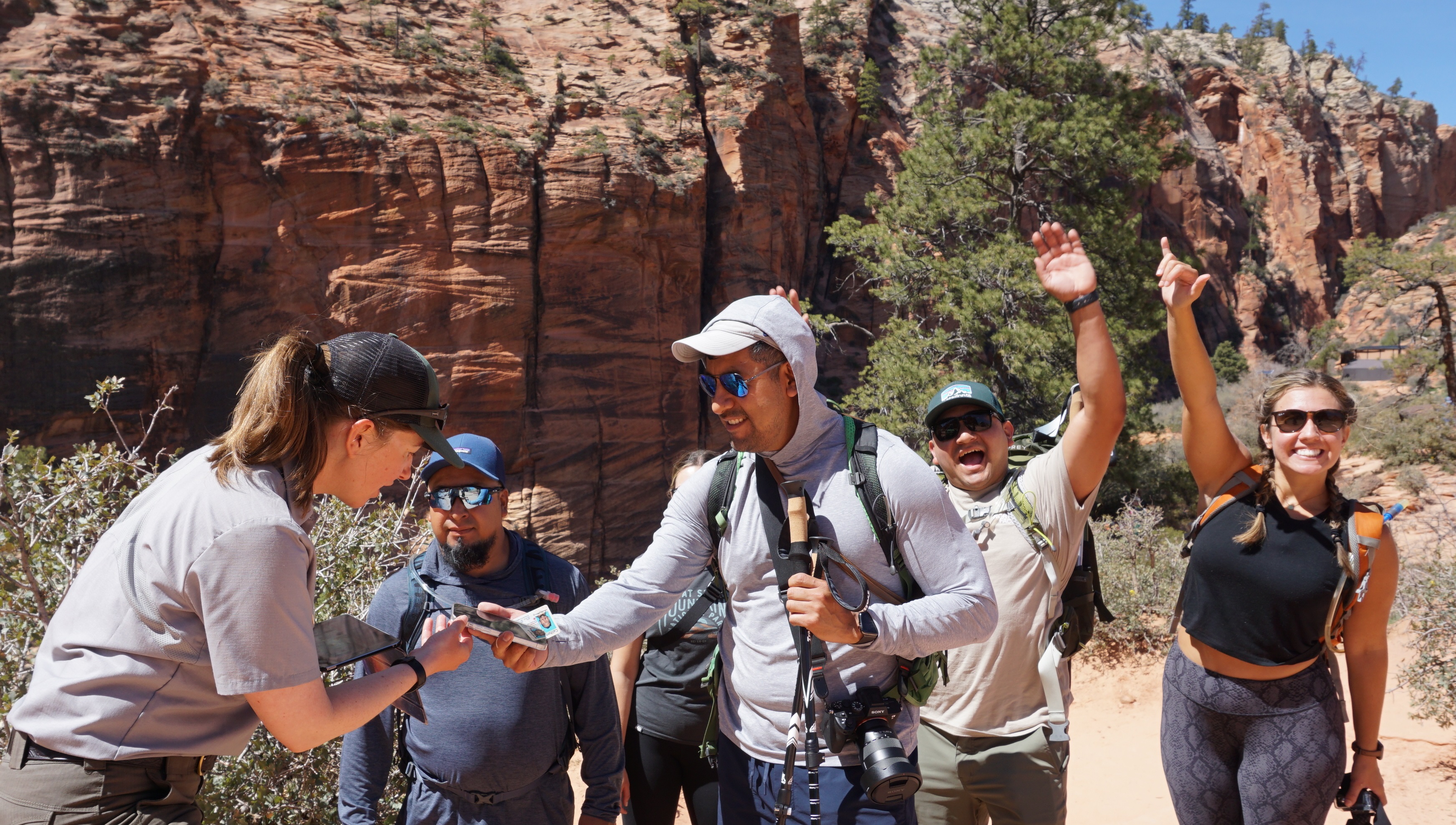 Park ranger checks Angels Landing permits in Zion National Park. A park ranger and several hikers stand at Scout Lookout. Two hikers have hands raised in celebration.