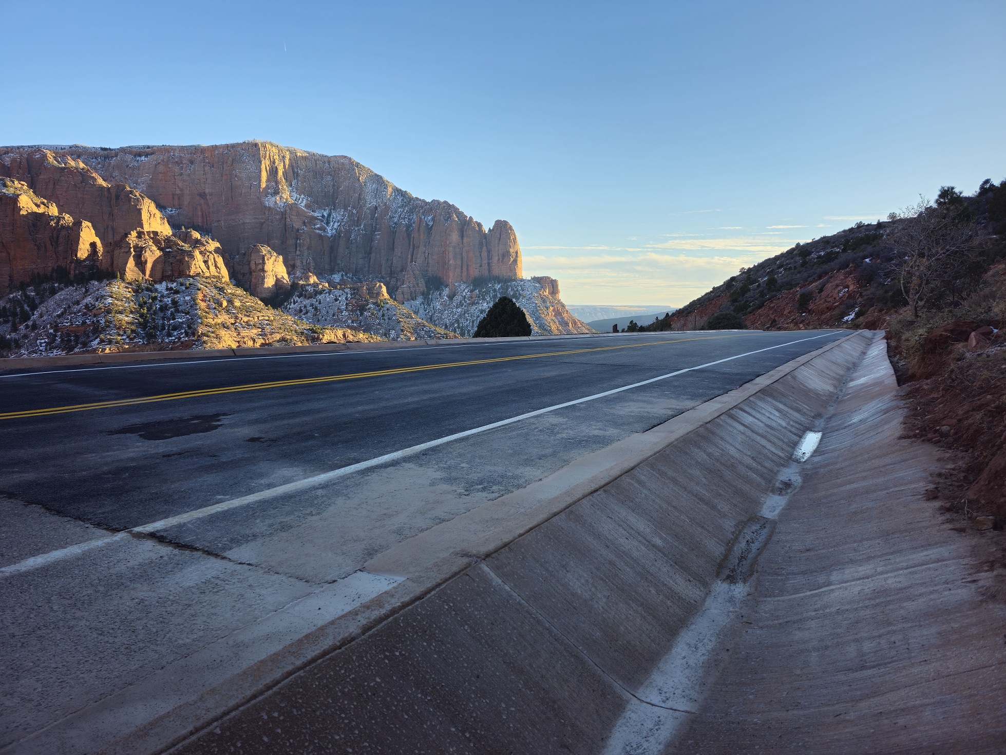 Newly paved drainage ditch next to newly paved and striped asphalt road with red rocks covered in show rising in the distance below a blue sky.