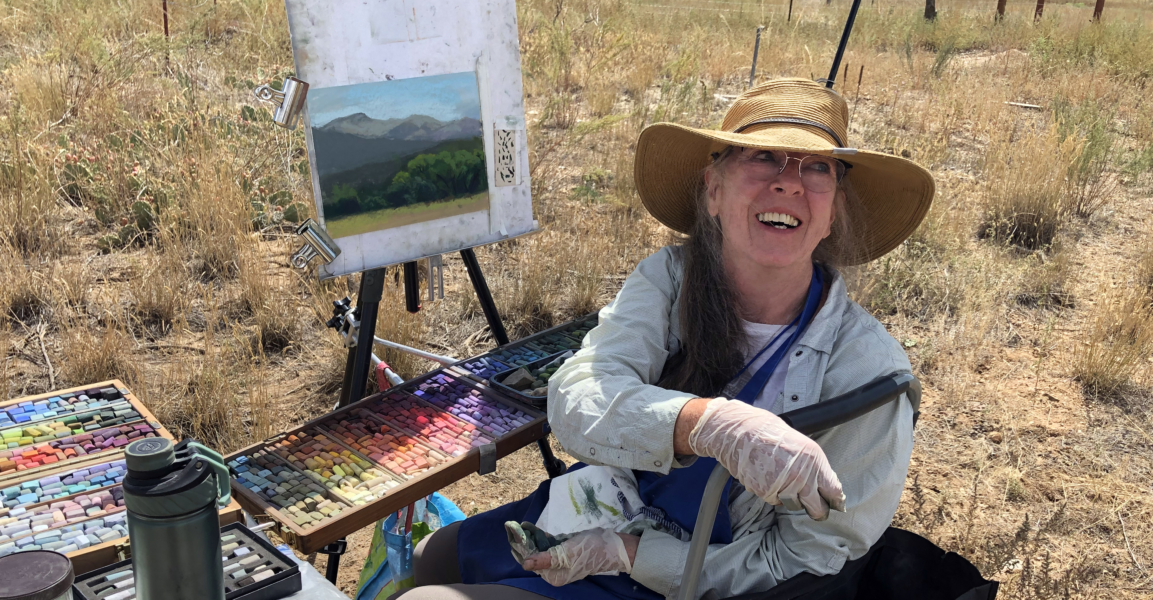 An artist wearing a hat sits next to a pastel piece she is working on.