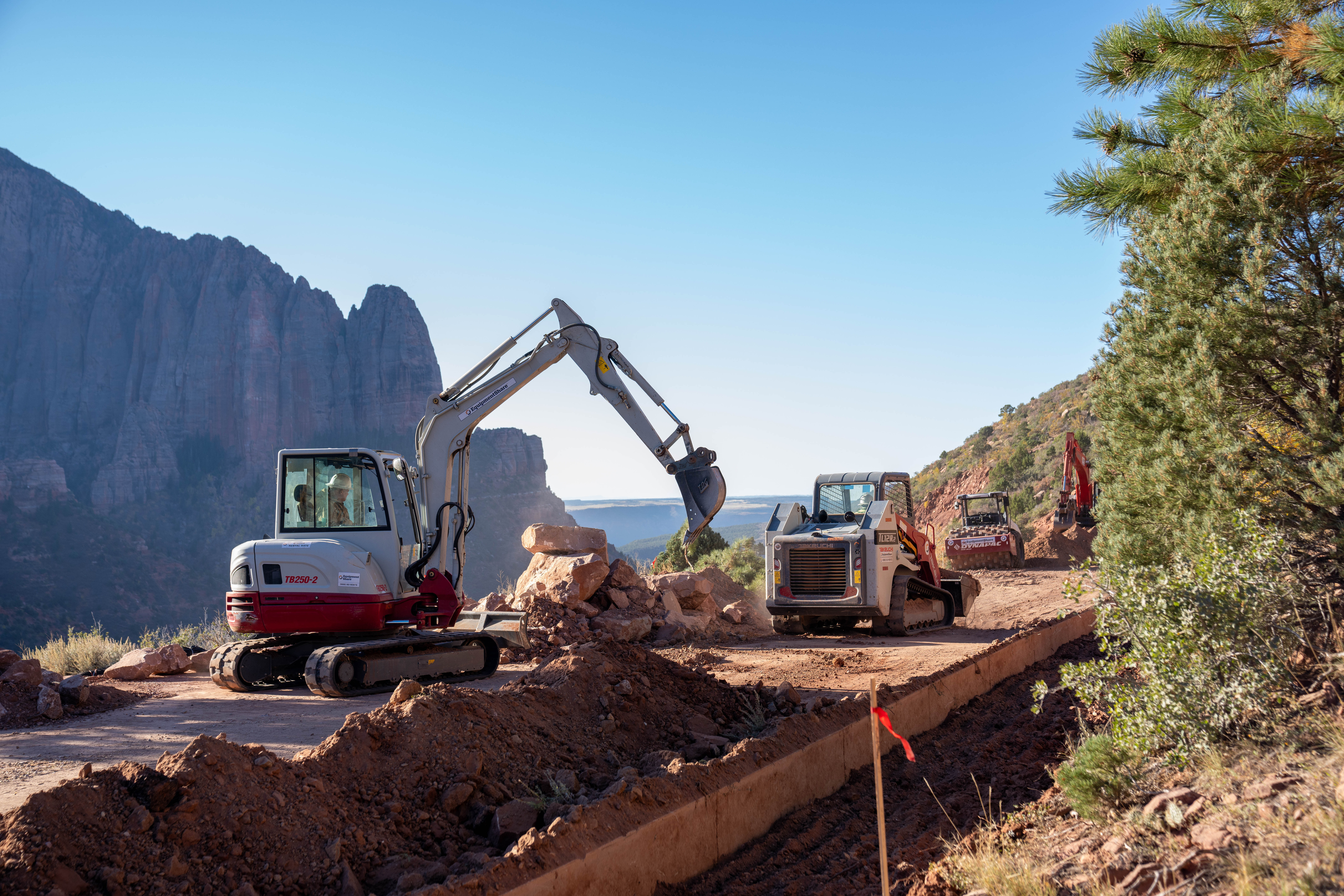 Excavating and other construction machinery sits on a partially graded dirt road-bed with concrete formwork in front of them and towering red rock and a clear sky behind.