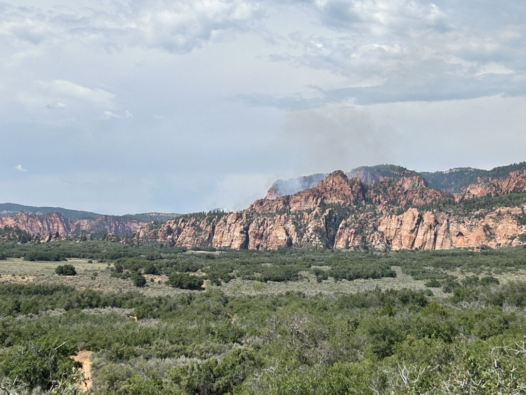 Hop Valley Fire July 24 2024 A green plain with red and orange sandstone cliffs in the background with smok coming from between the canyon