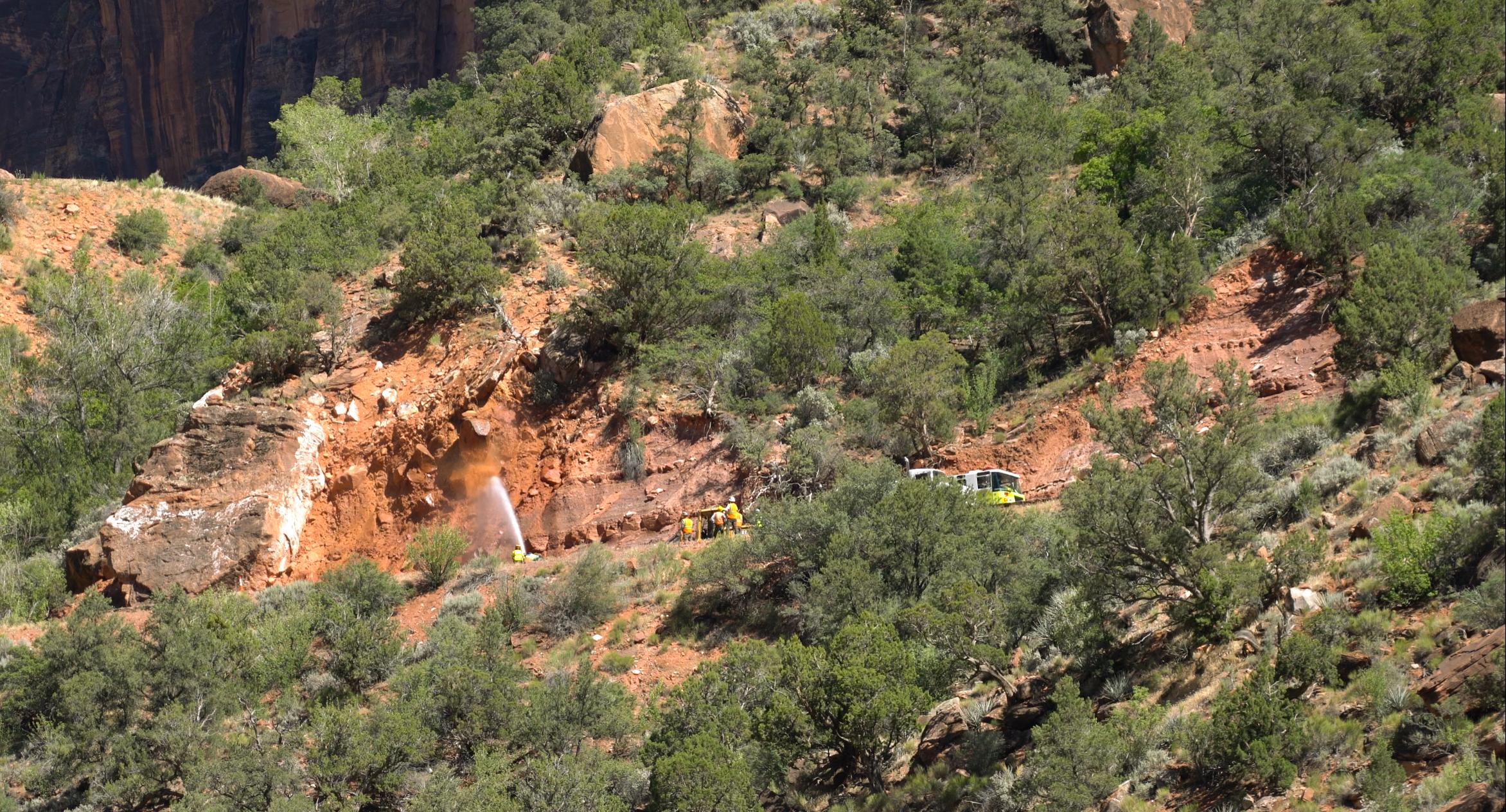 National Park Service staff dislodge loose rock above the Zion-Mt. Carmel Highway. National Park Service staff use water from a fire hose to dislodge loose rock above a road.