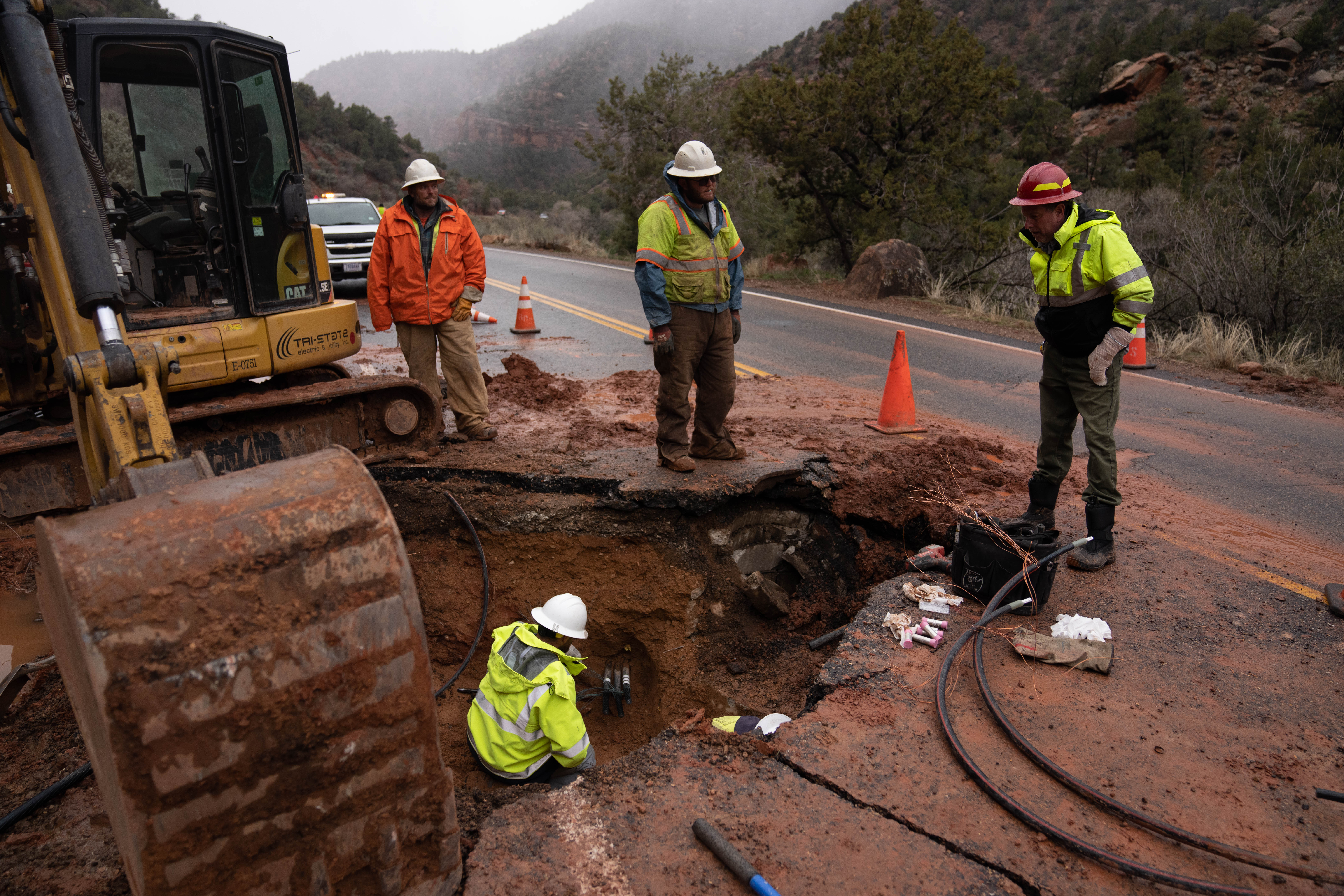 Four workers near a hole with heavy equipment to repair road damage resulting from a rockfall. Four people work near a large hole in Zion Canyon Scenic Drive. An excavator stands nearby as people work in and near a hole.