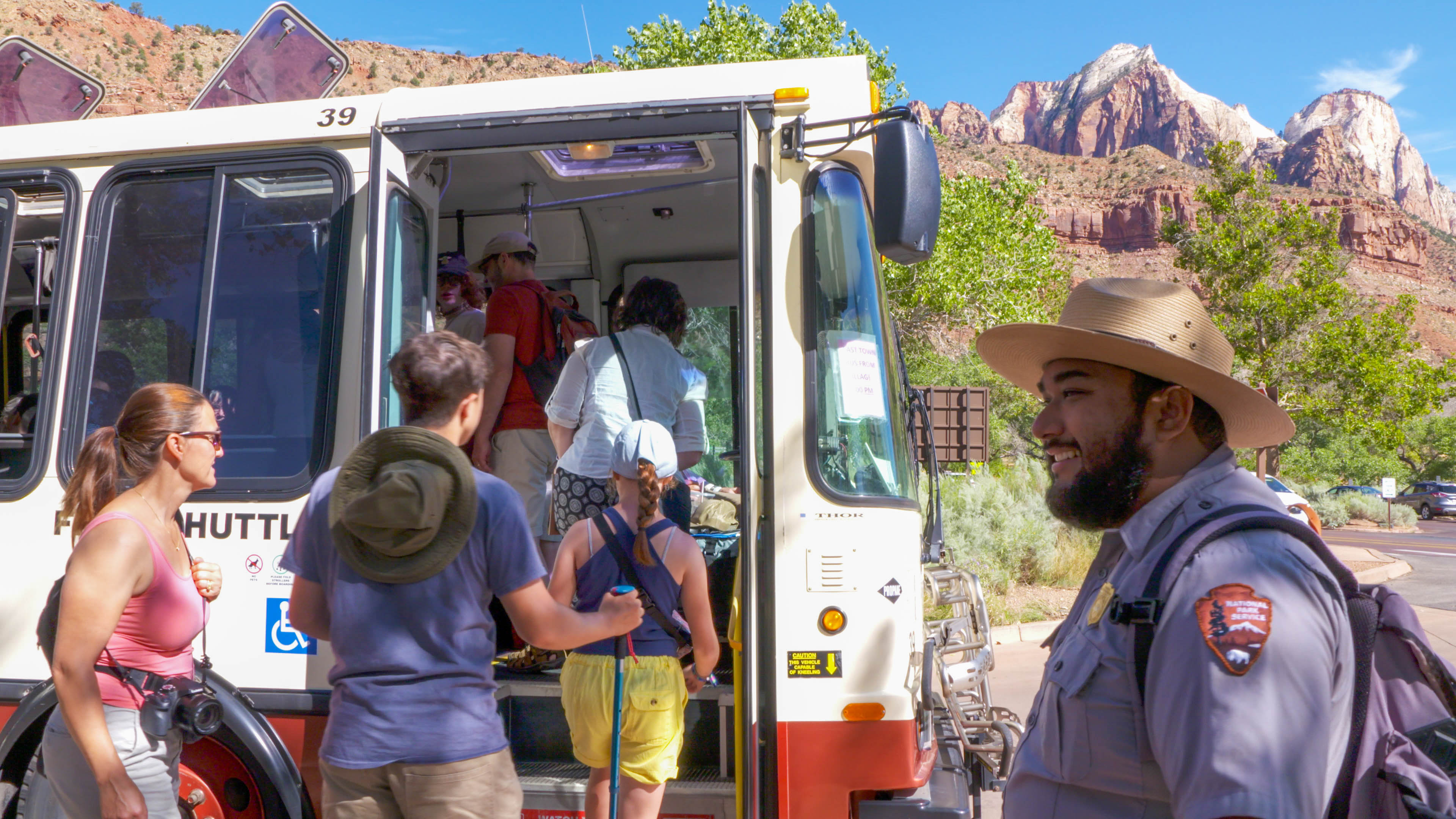 A ranger smiles as visitors board a Zion Cayon shuttle, with sandstone cliffs in the background