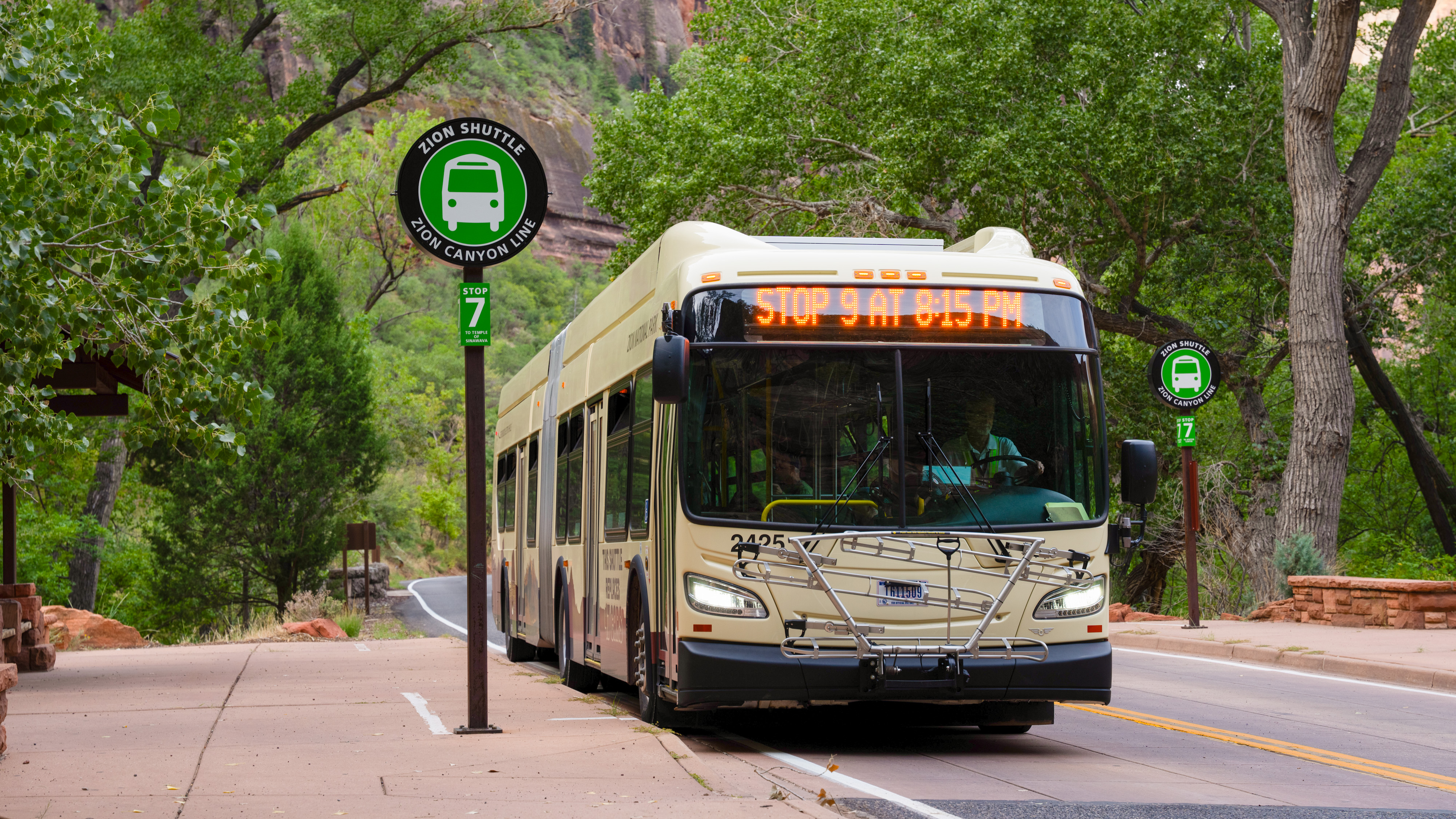 A white and orange shuttle bus stops at a small shelter along a canyon roadway.