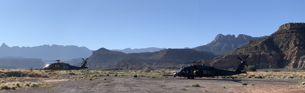 Two Blackhawk helicopters on the ground in a brushy wash, with canyon cliffs in the distance.