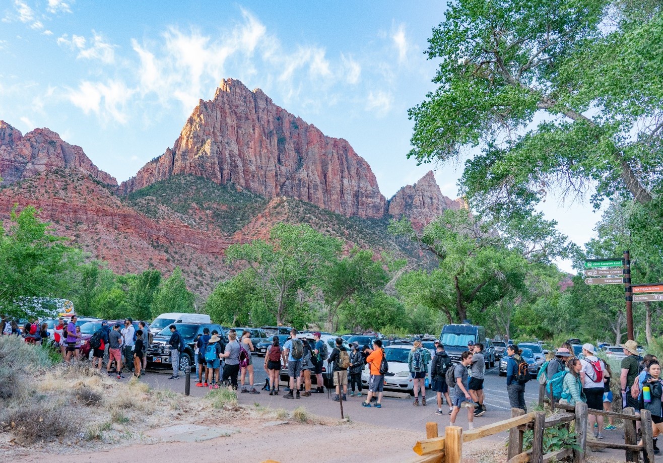 Zion Canyon Visitor Center shuttle line Memorial Day Weekend 2021 People stand in line near a parking lot with the red rock canyon wall of Zion National Park in the distance above them.