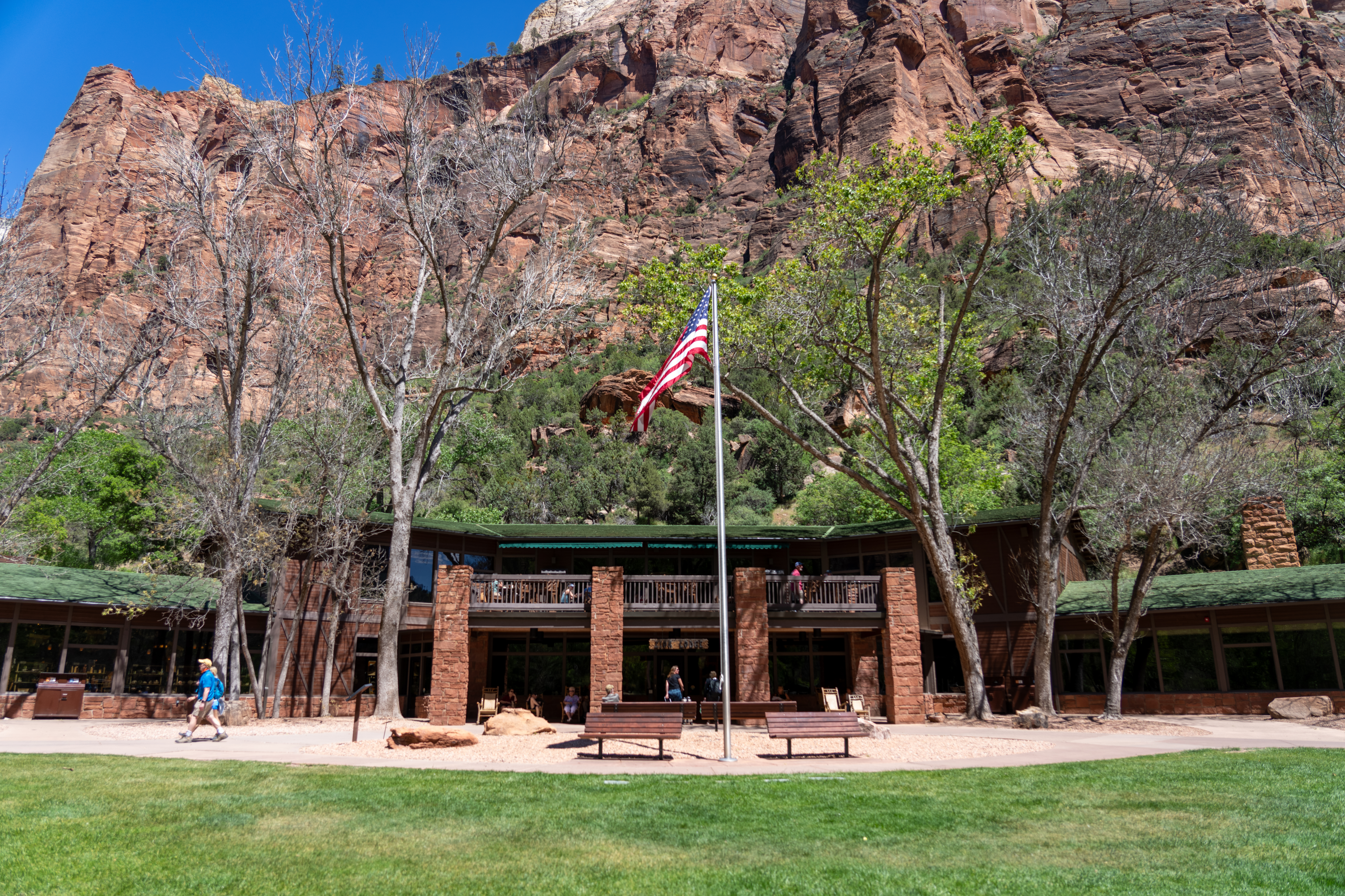 Zion Lodge_05132024_-1 Building with people standing outside, with a flag pole in teh foreground and sandstone cliffs and trees in teh background