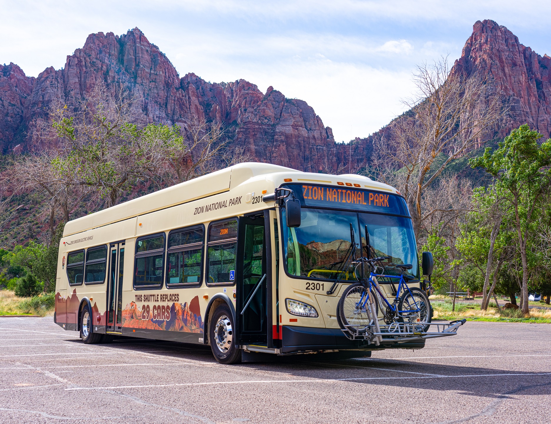 Electric shuttle bus with bikes on a rack mounted in front stands in Zion National Park with red rock towering behind. Text on bus reads, "This shuttle replaces 29 cars."