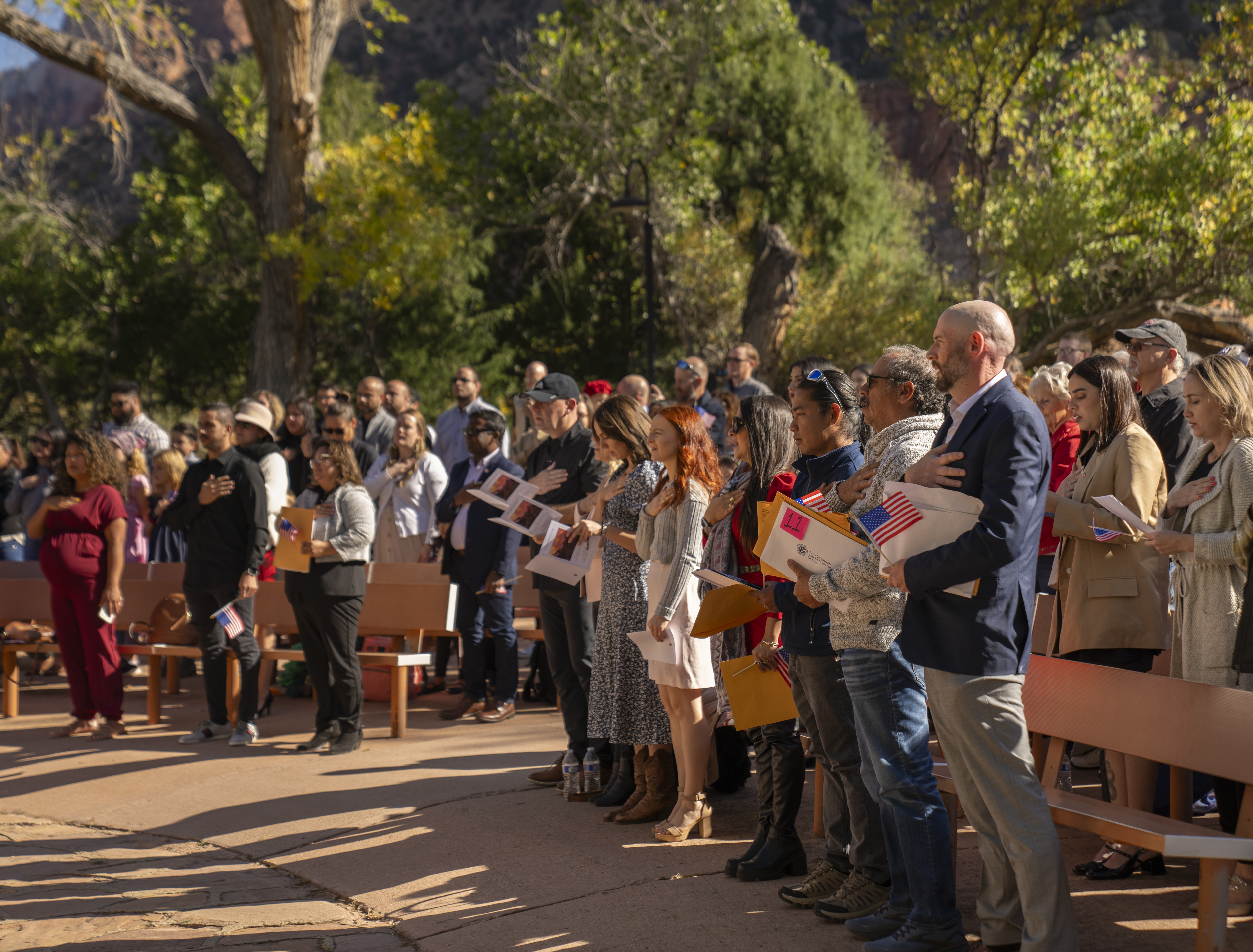 Zion Naturalization Ceremony 2023 People hold flags and put their hands over their heart for the national anthem with trees in the background.