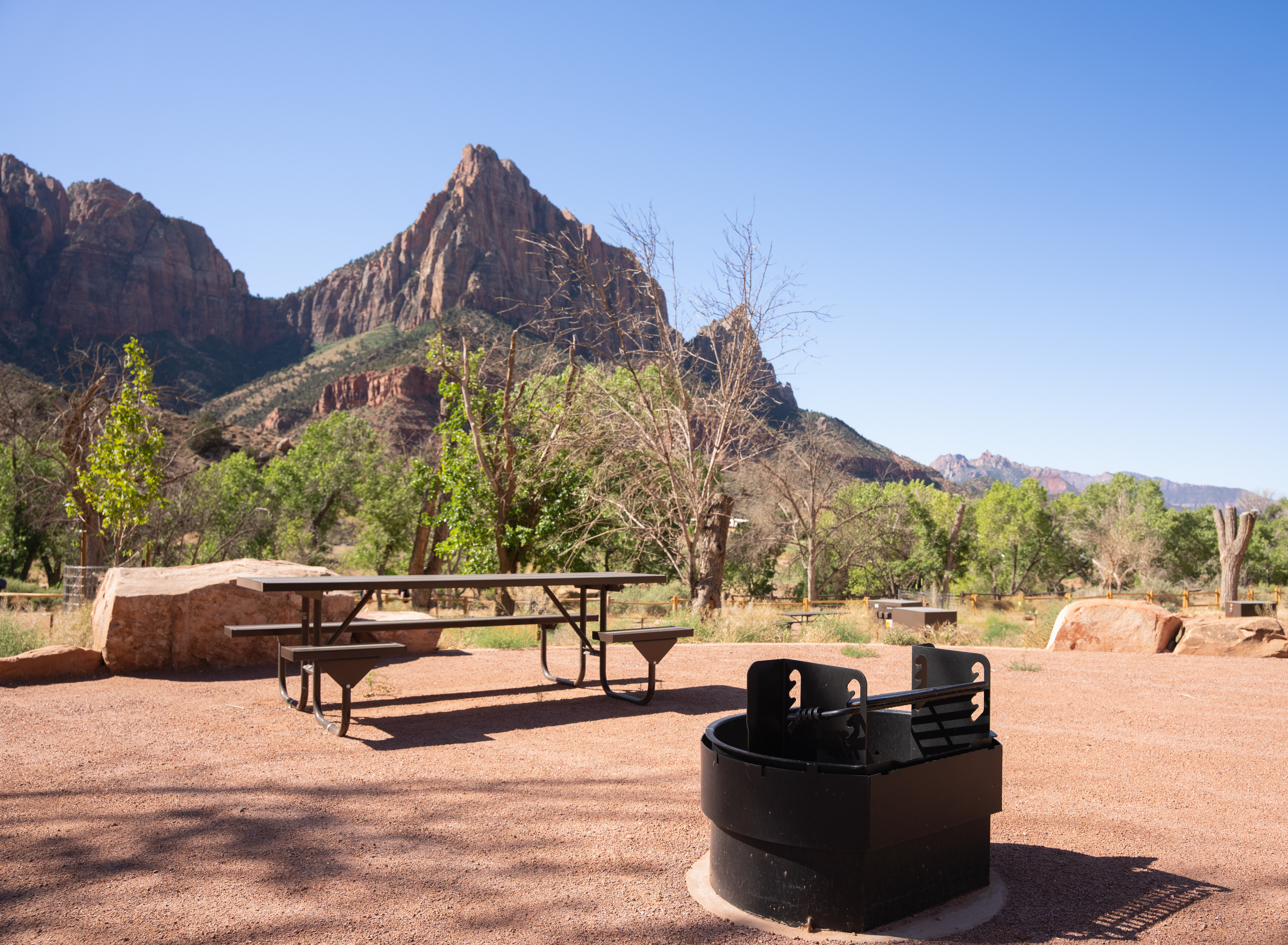 A picnic table and elevated fire pit on a flat surface. Green trees and a triangular sandstone formation are visible in the background.