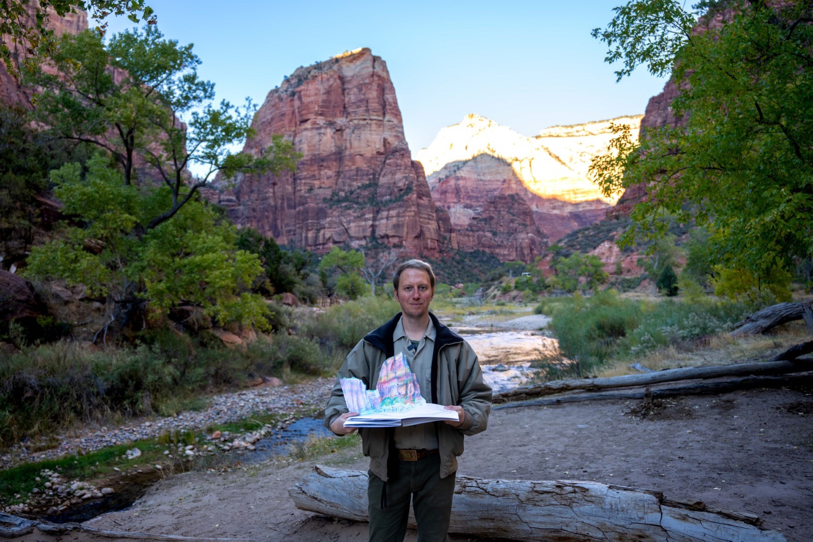 Fall 2024 Artist-in-Residence A man holding a popup book of a sandstone cliff standing in front of a sandstone cliff and river