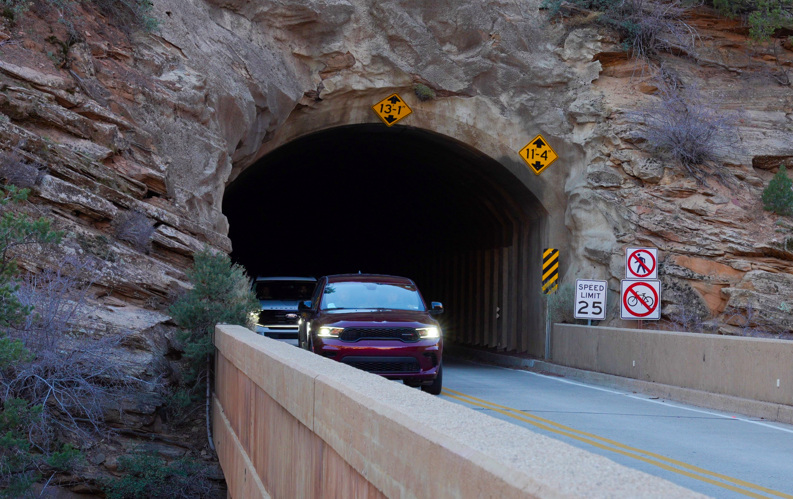 Two vehicles drive out of a large stone tunnel, which is surrounded by natural stone features.