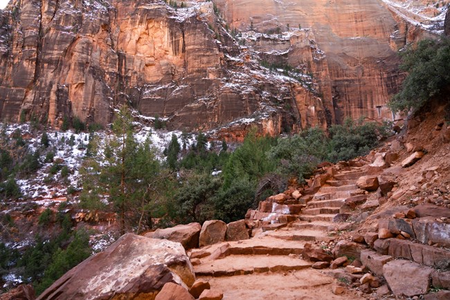 Steep stone steps curve along a cliffside and head towards a wall of snow-littered sandstone.