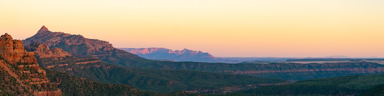 The sun sets over a vast wilderness. Green trees line every ridge and valley of the desert environment.