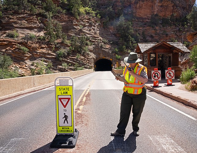 A ranger in a hi-vis vest talks into a radio on a road leading into a large tunnel carved into a cliff.