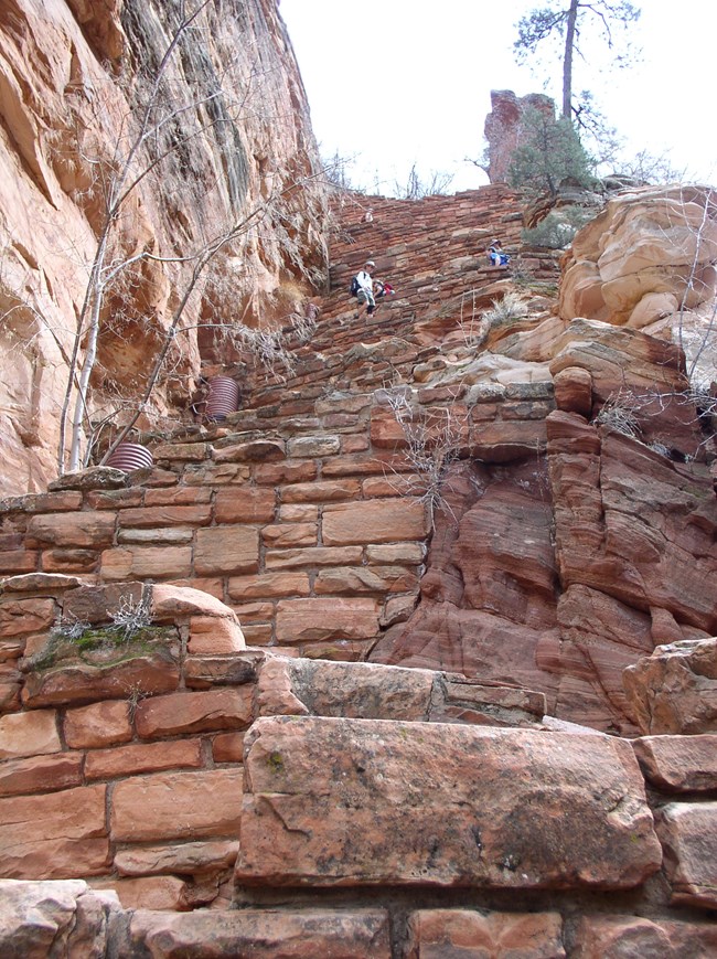 Hikers walk up narrow and steep switchbacks made of stone bricks and concrete.