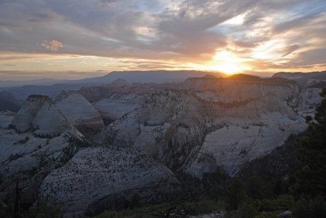 Sunset behind distant mountains, with a maze of canyons in the foreground.